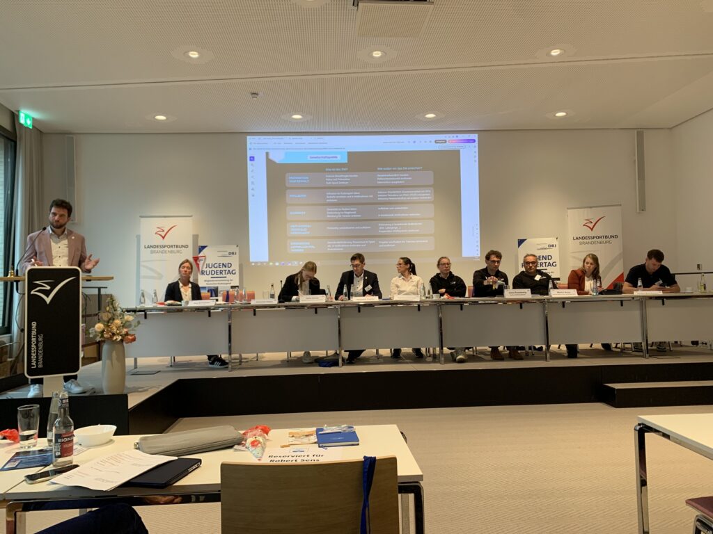 Der neue Vorstand der DRJ, am Rednerpult der wiedergewählte Vorsitzende Sebastian Haase und ganz links auf dem Podium die hauptamtliche Vorständin Jugend des DRV Sina Burmeister.