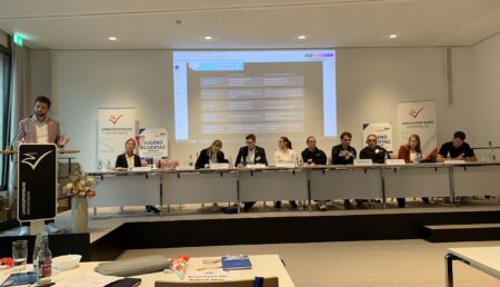 Der neue Vorstand der DRJ, am Rednerpult der wiedergewählte Vorsitzende Sebastian Haase und ganz links auf dem Podium die hauptamtliche Vorständin Jugend des DRV Sina Burmeister.